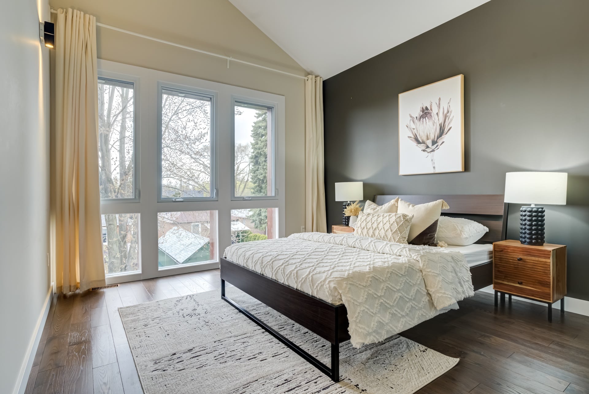 Staged bedroom with large windows, hardwood floors, area rug, bed with white comforter, and modern lamps for real estate appeal.