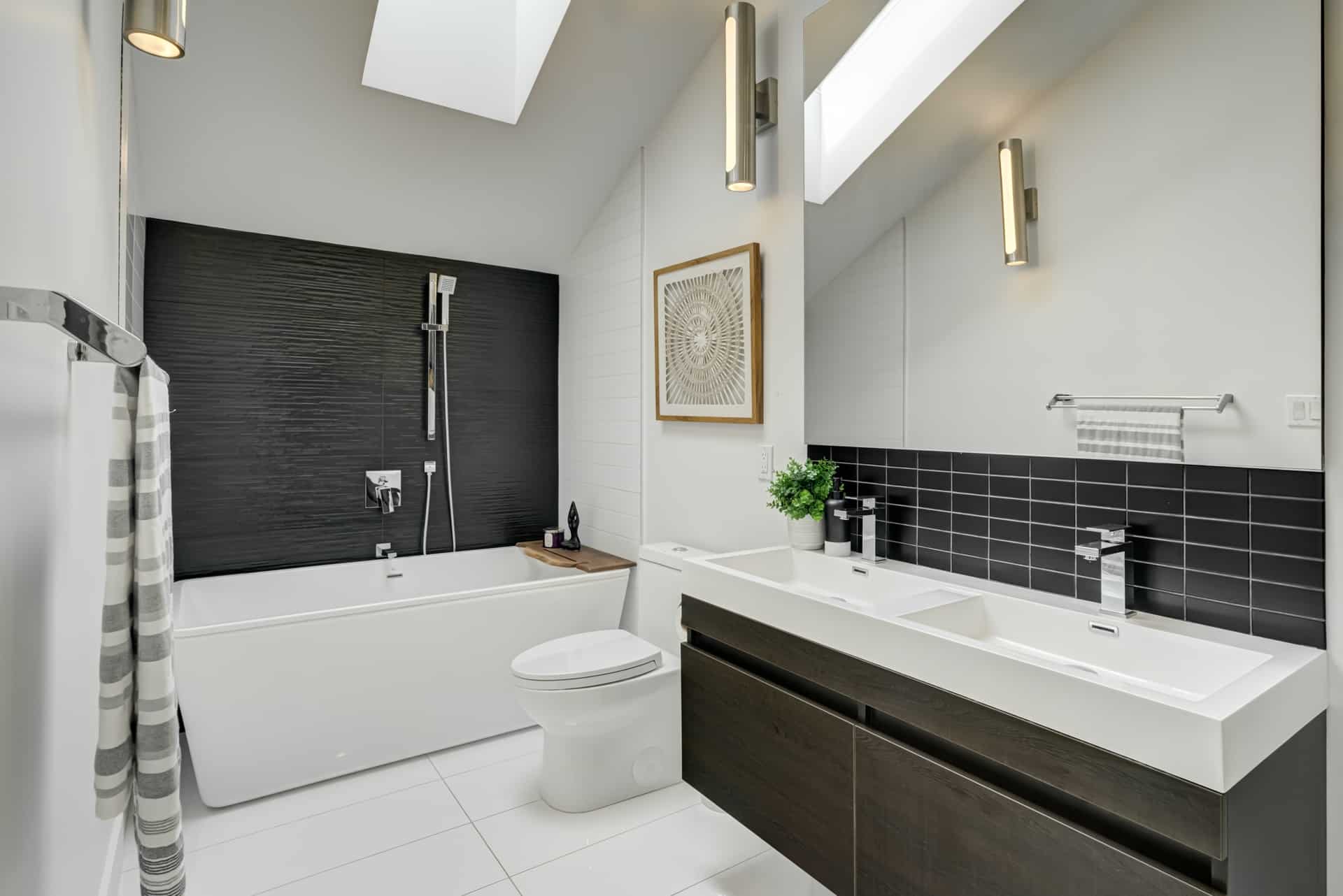 Modern bathroom staging with white tub, sleek vanity, black tile accent, and skylights for bright, stylish home decor.